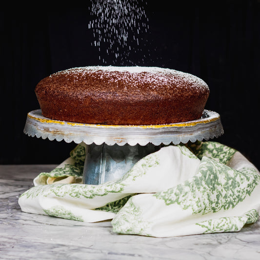 Chocolate Lemon Olive Oil Cake recipe plated on silver cake stand being sprinkles with powdered sugar