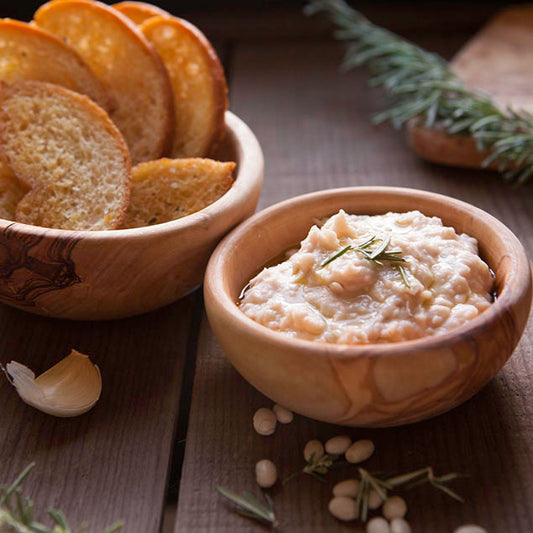 Pasolivo Rosemary White Bean Dip Recipe on a table with a bowl of crostini bread and garlic cloves